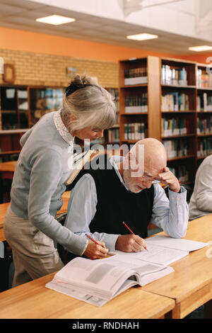 Female professor checking the work of an elderly student in classroom. Senior man writing notes in his book while a senior lecturer guides him. Stock Photo