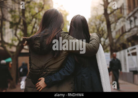 Two women walking together on street holding each other. Rear view of women in winter clothes walking on street with arms around each other. Stock Photo