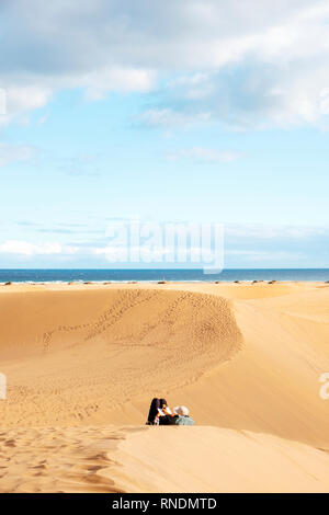 a view of the sand dunes of Maspalomas, in the Canary Islands, Spain, with an unrecognizable woman resting in the foreground and the Atlantic ocean in Stock Photo