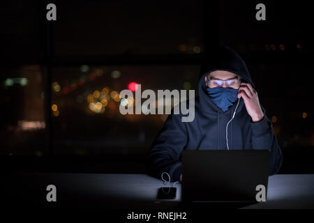 Young talented hacker using laptop computer while working in dark office with big city lights in the background at night Stock Photo