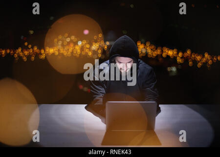 Young talented hacker using laptop computer while working in dark office with big city lights in the background at night Stock Photo