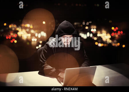 Young talented hacker using laptop computer while working in dark office with big city lights in the background at night Stock Photo