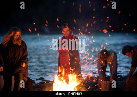 Mid-adult man with a blanket around him as he stands beside a bonfire with people on a rocky beach Stock Photo