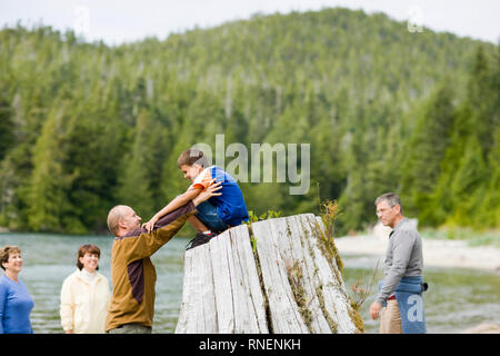 Young boy atop a large tree stump being helped down by his mid-adult father beside a lake.