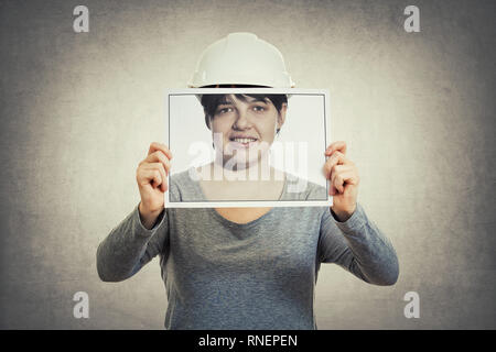 Young woman engineer wearing protective helmet, holding a paper photo covering her face, like a mask for hiding the true emotion. Worker private life, Stock Photo