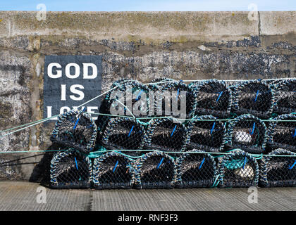 Creel nets piled up on a sunny day with a "God is Love" sign painted on the harbour wall behind them, Gardenstown, Aberdeenshire Stock Photo
