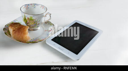 On a white surface we have a small plate and a porcelain cup, a half croissant and an electronic tablet. Copy space at right and hi key. Stock Photo