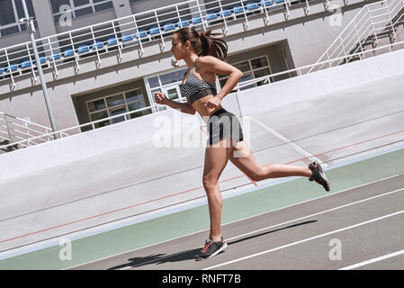 Young woman wearing black sportswear running on racetrack during training session. Female runner practicing on athletics race track. She is concentrated and motivated. Side view Stock Photo