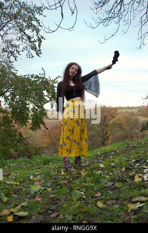 A brunette caucasian lady with long lace black sleeves and a fashionable floral outfit holds a leaf and sticks out her tongue, standing on a hill in a Stock Photo
