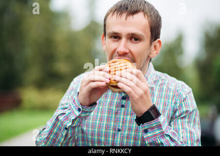 Man in checked shirt eating tasty burger outdoors. Stock Photo