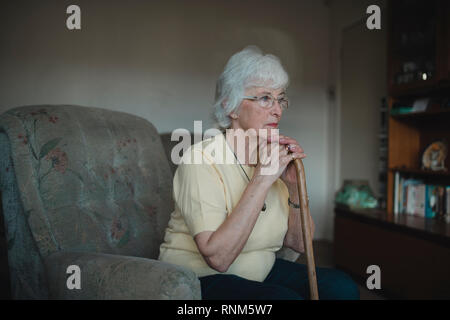 Senior woman is looking sad while sitting alone on the sofa in her living room. She is resting on her walking stick. Stock Photo