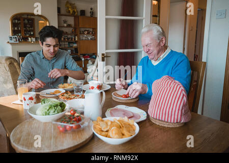 Senior man is having lunch at home with his grandson. They are eating sandwiches, pizza and potato chips. Stock Photo