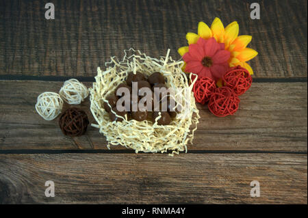 Round chocolates in a nest of straw, multicolored decorative balls and flower buds on a wooden table. Close-up. Stock Photo