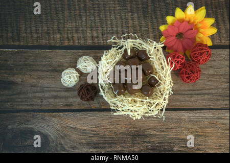 Round chocolates in a nest of straw, multicolored decorative balls and flower buds on a wooden table. Close-up. Stock Photo