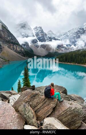 Young woman sitting in front of a lake looking into mountain landscape, clouds hanging between mountain peaks, reflection in turquoise lake, Moraine L Stock Photo