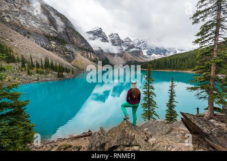 Young woman standing in front of a lake looking into mountain scenery, clouds hanging between mountain peaks, reflection in turquoise lake, Moraine La Stock Photo