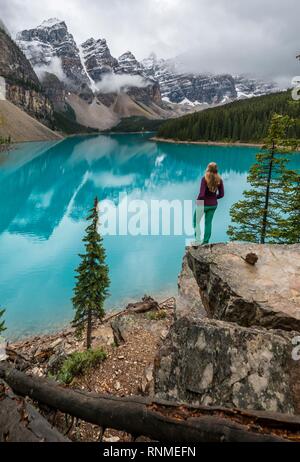 Young woman standing in front of a lake looking into mountain scenery, clouds hanging between mountain peaks, reflection in turquoise lake, Moraine La Stock Photo