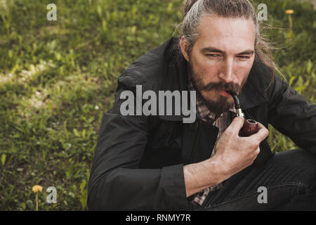 Serious bearded man smoking pipe in summer park, close up portrait Stock Photo