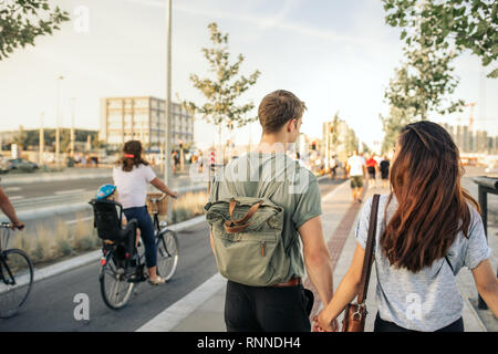 Rearview of a young couple walking hand in hand together along a city sidewalk in the afternoon Stock Photo