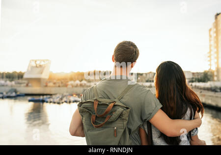Rearview of a young couple standing arm in arm together looking out over a harbor in the late afternoon Stock Photo