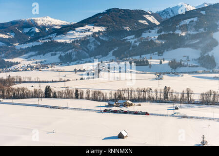 Red and white train ÖBB passing the snow-covered fields in a scenic winter mountain landscape, Dachstein massif, Liezen District, Styria, Austria Stock Photo