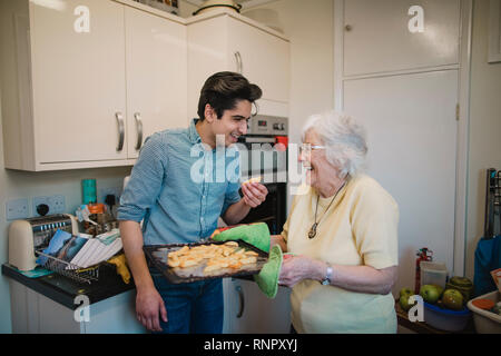 Teenage boy and his grandmother are laughing in the kitchen together after the teenager has taken a potato chip from the tray his grandmother has remo Stock Photo