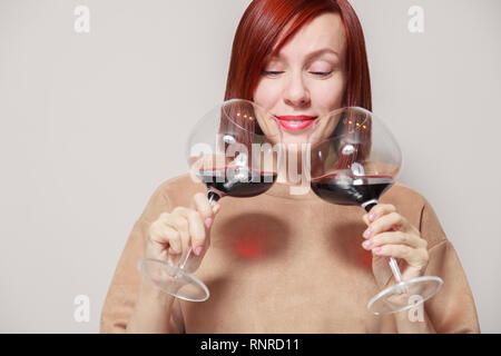 Young cheerful redhaired girl holds and sniffs two glasses of red wine and assesses the aroma and notes at professional blind tasting. Concept alcohol Stock Photo