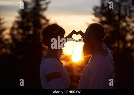 Silhouettes of romantic couple in love in the evening. Bearded man and happy woman making a heart with hands, smiling and looking at each. Amazing sunset, cloudy sky and forest on blurred background Stock Photo