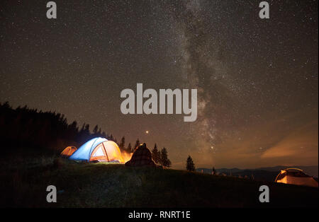 Night camping in mountains near forest. Bach view couple hikers covered by blanket plaid resting together, sitting beside campfire and tourist tent under night starry sky full of stars and Milky way. Stock Photo