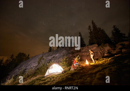Night summer camping in the mountains. Happy couple travellers having a rest together beside campfire and illuminated tourist tent. On background big boulder, forest and beautiful night starry sky. Stock Photo