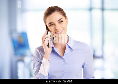 Portrait shot of young businesswoman using her mobile phone while standing in the office and talking with somebody. Stock Photo