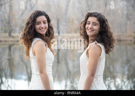 Side view of cheerful twin sisters standing at calm lake smiling and ...