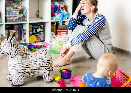 Tired of everyday household mother sitting on floor with hands on face. Kid playing in messy room. Scaterred toys and disorder. Happy parenting. Stock Photo