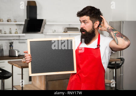 What to cook. urprise. This is what you need. Hipster in kitchen. Restaurant or cafe menu. Advertisement. Bearded man in red apron. Mature male. Bearded man cook. Man chef with board, copy space. Stock Photo