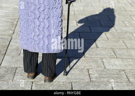 Old woman walking with a cane, long shadow on pavement. Concept for old age, diseases of the spine, elderly or blind person Stock Photo