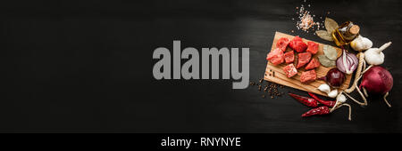 Uncooked fresh diced beef meat with herbs and oil on an old rustic wooden kitchen board over black background. Top view with free space for text Stock Photo