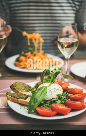 Italian dinner at bistrot with caprese salad with mozzarella, tomatoes, basil, pesto toasts, spaghetti pasta, white wine. Man eating pasta, looking in Stock Photo