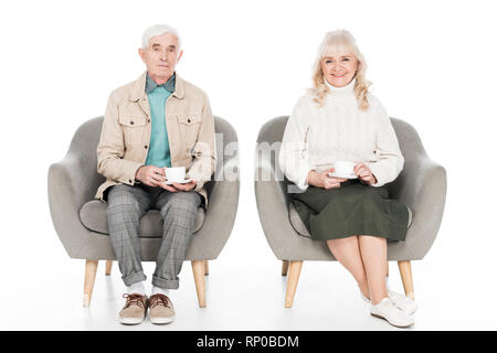happy retired husband and wife holding cups with tea while sitting in armchairs isolated on white Stock Photo