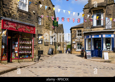 Traditional sweet shop, historic inns, Tourist Information & bunting over cobbled road - Main Street, Haworth village, West Yorkshire, England, UK. Stock Photo