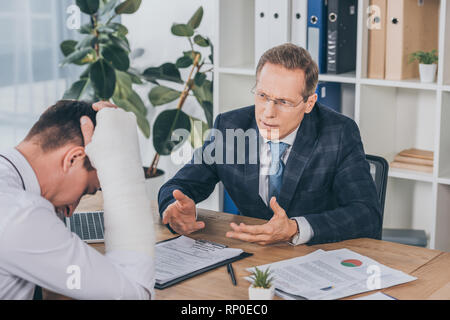 worker with broken arm sitting at table and reading documents opposite to businessman in blue jacket in office, compensation concept Stock Photo