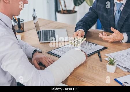 cropped view of businessman in blue jacket sitting at table and giving money to worker with broken arm in office, compensation concept Stock Photo