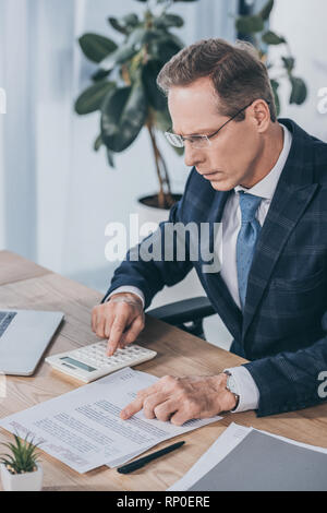 middle aged businessman sitting at table, reading document and counting with calculator in office, compensation concept Stock Photo