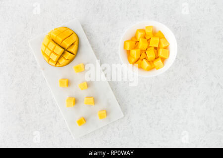 Top view of cut mango half and mango cubes on white marble cutting board and in white bowl. Stock Photo