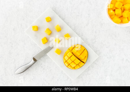 Top view of cut mango half and mango cubes on white marble cutting board with knife and in white bowl. Stock Photo