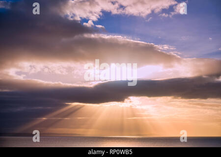 Rays of sun are shining through dark and moody clouds reflecting on the sea. Stock Photo