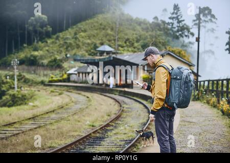 Young tourist with backpack using phone and waiting for train. Mountains railway station in clouds, Sri Lanka. Stock Photo