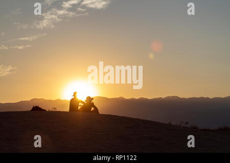 A couple sitting on a sand dune at sunset silhouetted against the sun, Mesquite Flat Sand Dunes, Death Valley National Park, California, United States Stock Photo