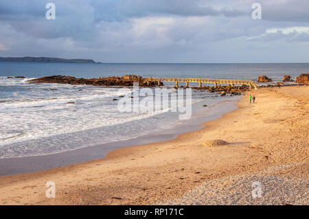 Ballycastle, Northern Ireland, UK. Atlantic coast. Beach with bridge, island, rocks, waves and unrecognizable waking pair in sunset light with dark he Stock Photo