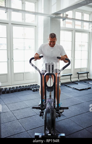 Mature man in sportswear riding a stationary bike pushing himself to his limit while working out alone at the gym Stock Photo