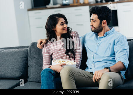 attractive latin woman holding bowl with popcorn and looking at husband at home Stock Photo